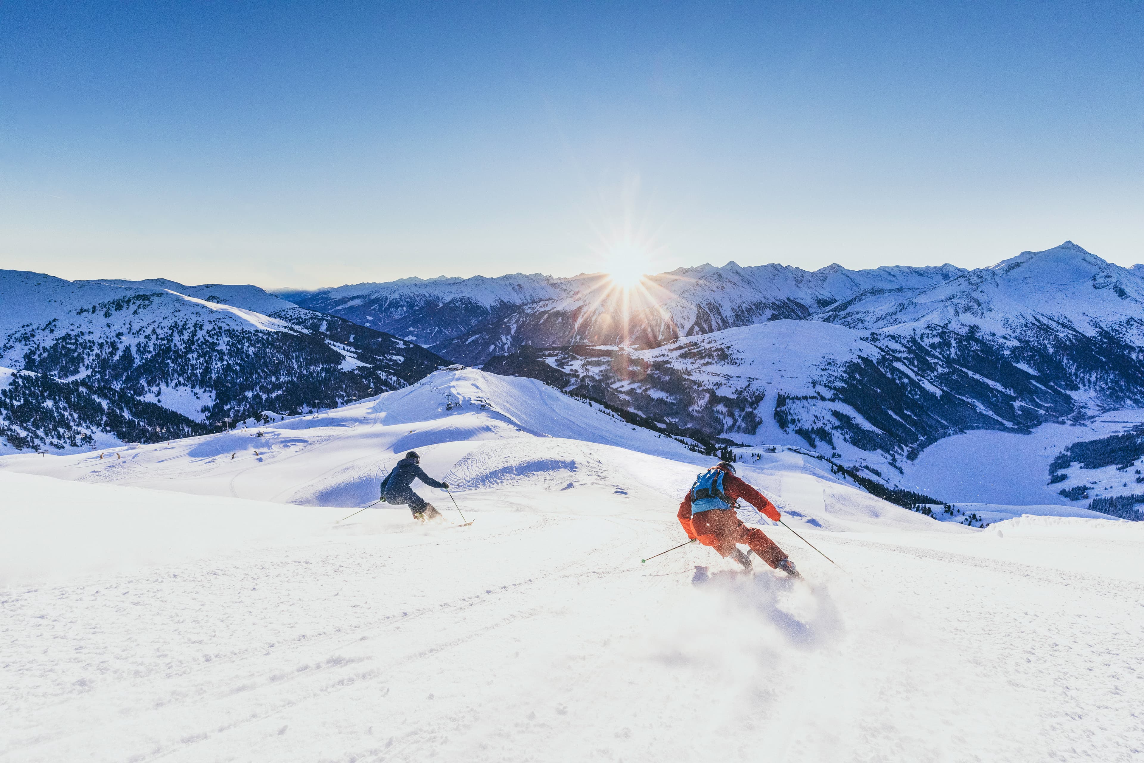 Skiiers skiing down mountain piste at sunset